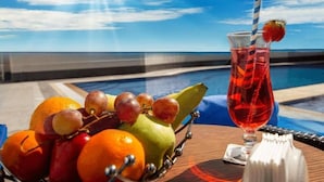 A fruit bowl with oranges, apples, and grapes, and a glass of red drink with a straw and a strawberry on a table by a pool with a clear view of the ocean. - Studio Apartment in Al Qusais With Free Breakfast (Dubai)