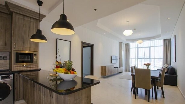 A modern kitchen with a central island, a dining area with a table and chairs, and a large window providing natural light. - One Bedroom Apartment in Al Qusais By E R (Dubai)