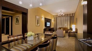 Camera d'albergo con zona pranzo, un tavolo con piano in vetro e sedie imbottite a righe, una televisione, un divano e una lampada.