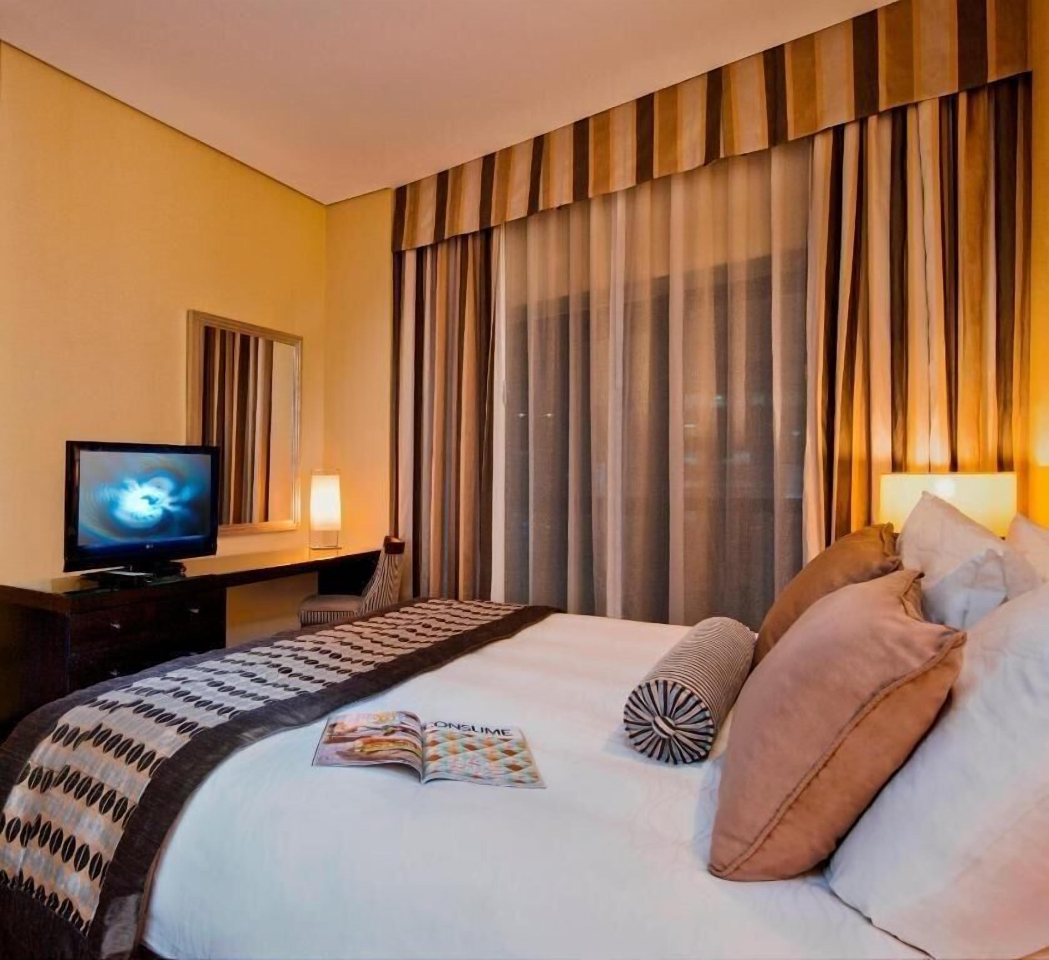 A hotel room with a bed, a television, a desk, and a window with curtains.