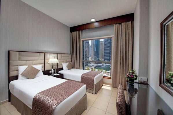 A hotel room with two beds, a large window with a city view, a desk with a lamp, and a small table with a vase of flowers.