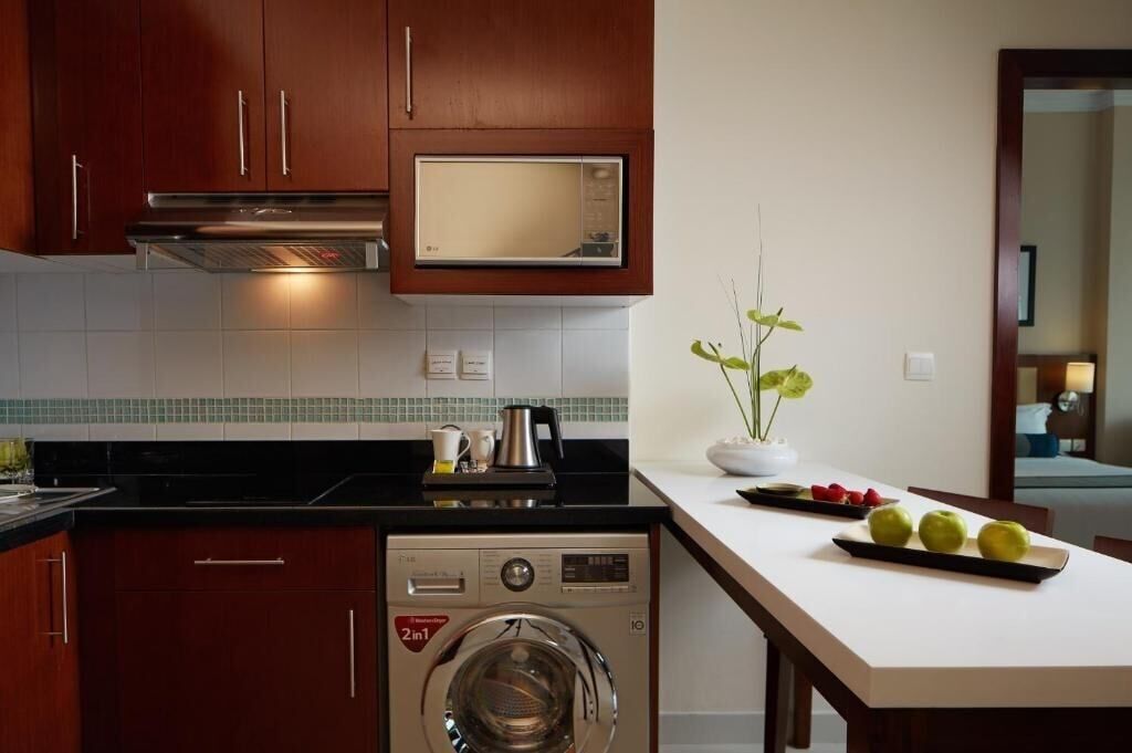 A compact kitchen with wooden cabinets, a built-in microwave, a washing machine, and a dining area with a table and chairs.