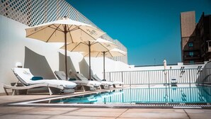 A rooftop pool area with sun loungers and umbrellas, surrounded by a white fence and buildings. - One Bedroom Apartment In Jaddaf By E R (Dubai)