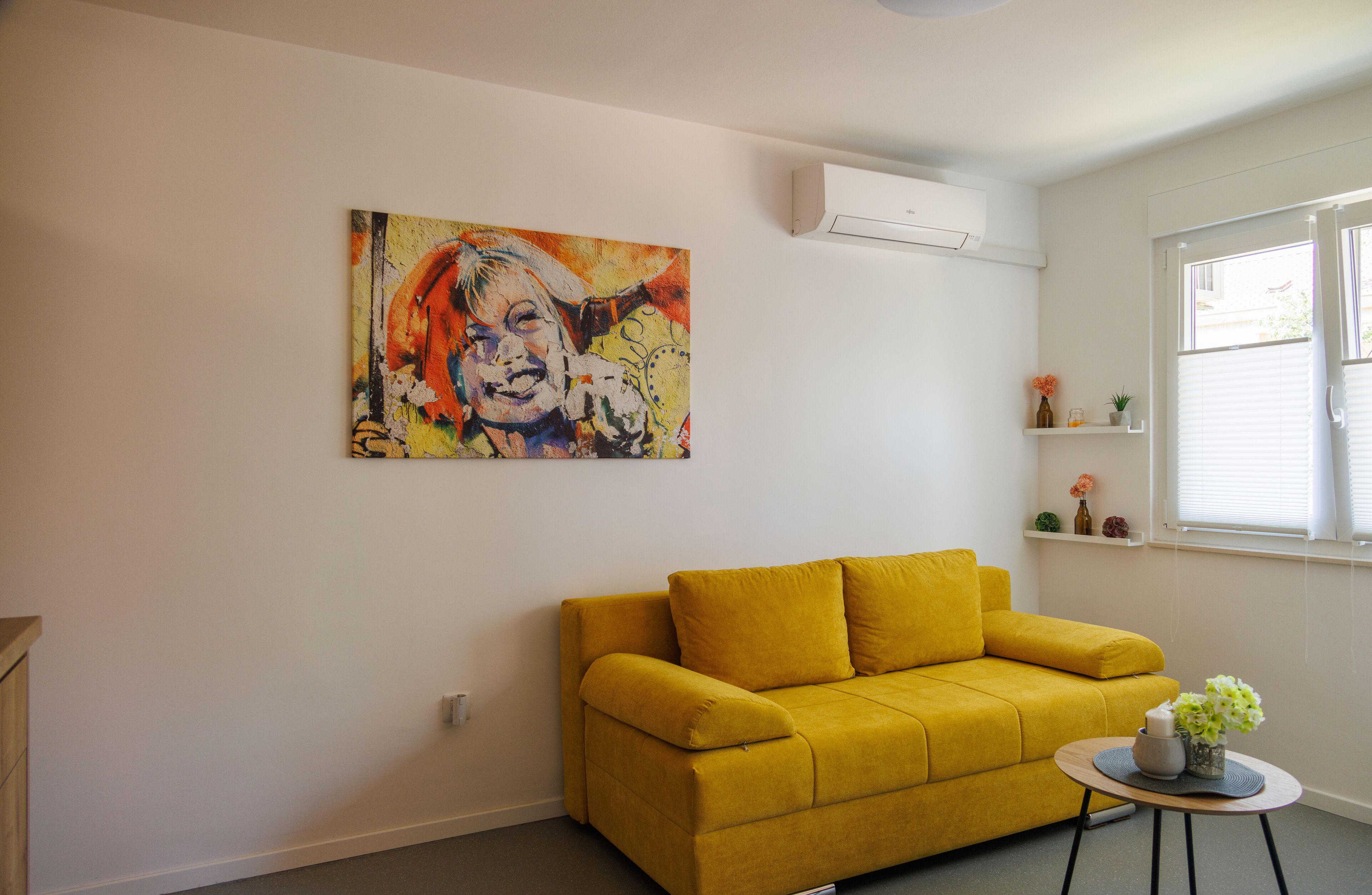 A living room with a yellow sofa, a painting on the wall, and a small table with a vase.