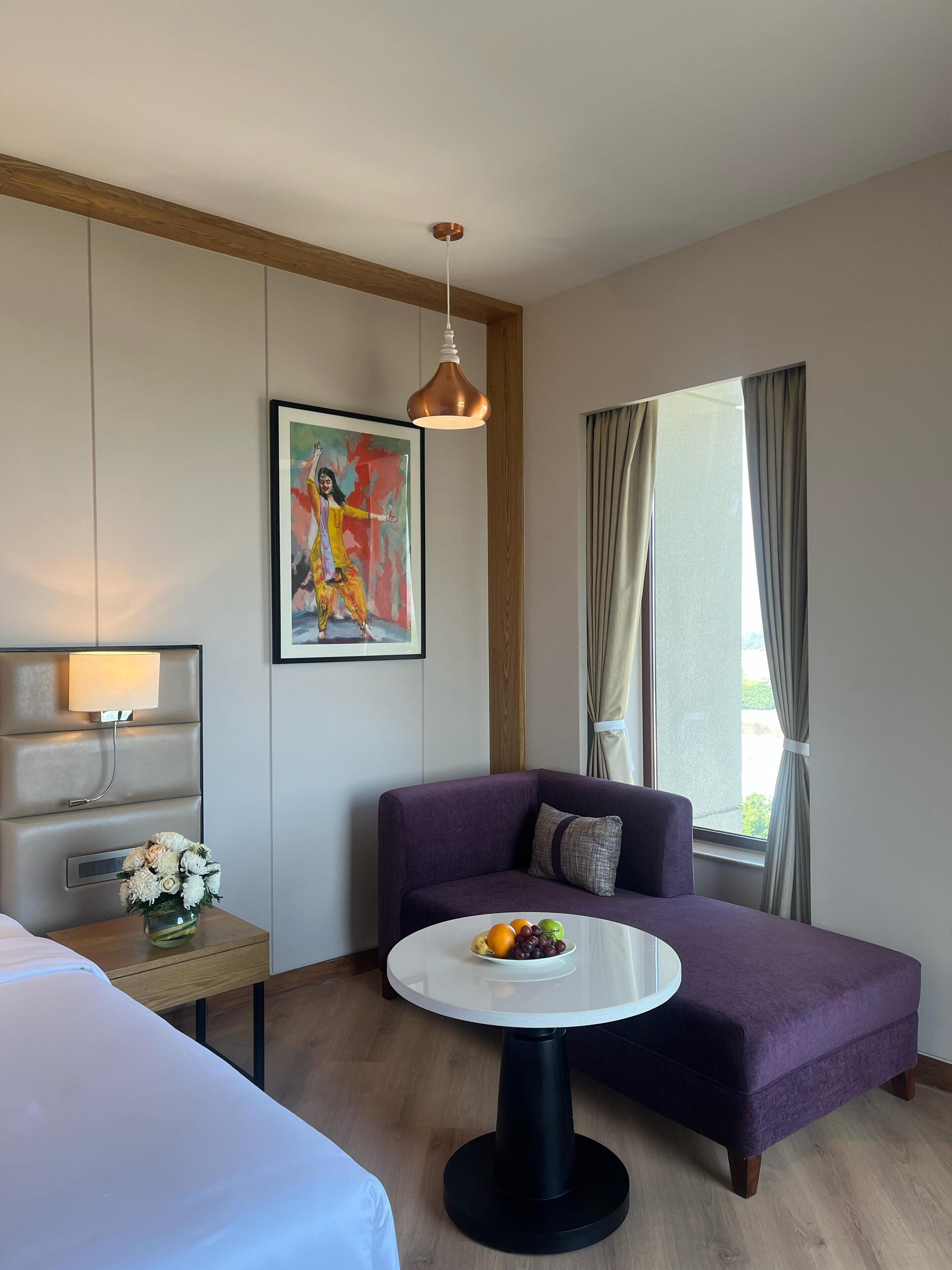 A hotel room with a purple sofa, a round table with fruit, a painting on the wall, and a window with curtains.