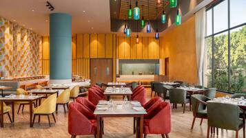 A modern restaurant with wooden flooring, red and green chairs, and a large window offering a view of trees.