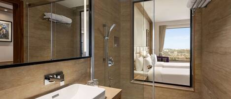 Un baño moderno con un espejo grande, un lavabo blanco, una ducha con mampara de cristal y vistas a un dormitorio con cama y ventana.
