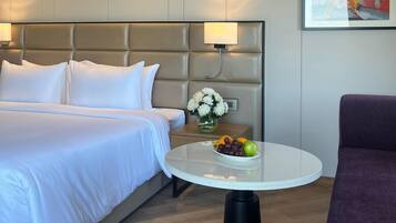 A hotel room with a bed, a round table with fruit, a purple chair, and a painting on the wall.