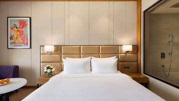 Habitación de hotel con una cama grande, cabecera con iluminación integrada, mesita de noche con un jarrón de flores y zona de ducha.