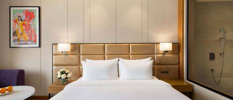 A hotel room with a large bed, a headboard with integrated lighting, a nightstand with a vase of flowers, and a shower area.