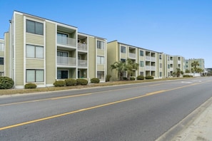 A row of multi-story apartment buildings with balconies, a clear sky, and a wide road. - Cozy 2BR North Myrtle Beach Condo—Walk to Beach, Shops & Dining (North Myrtle Beach)