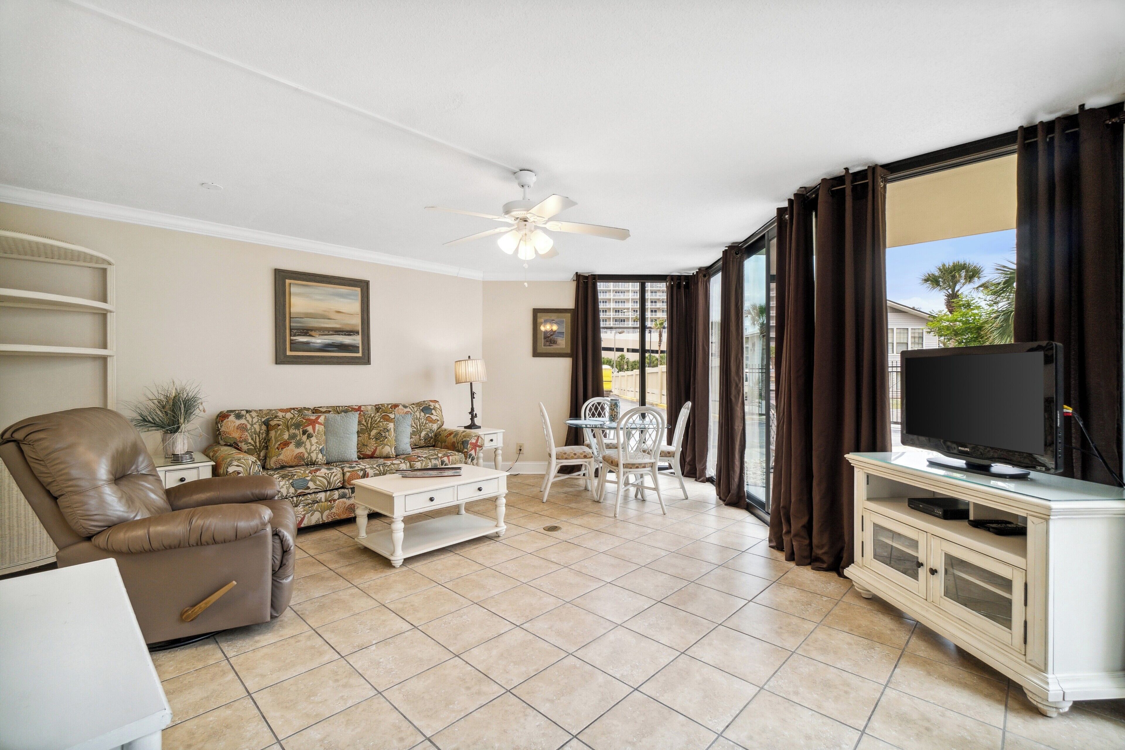 A living room with a sofa, a recliner, a coffee table, a TV on a cabinet, and a dining area with chairs and a table.