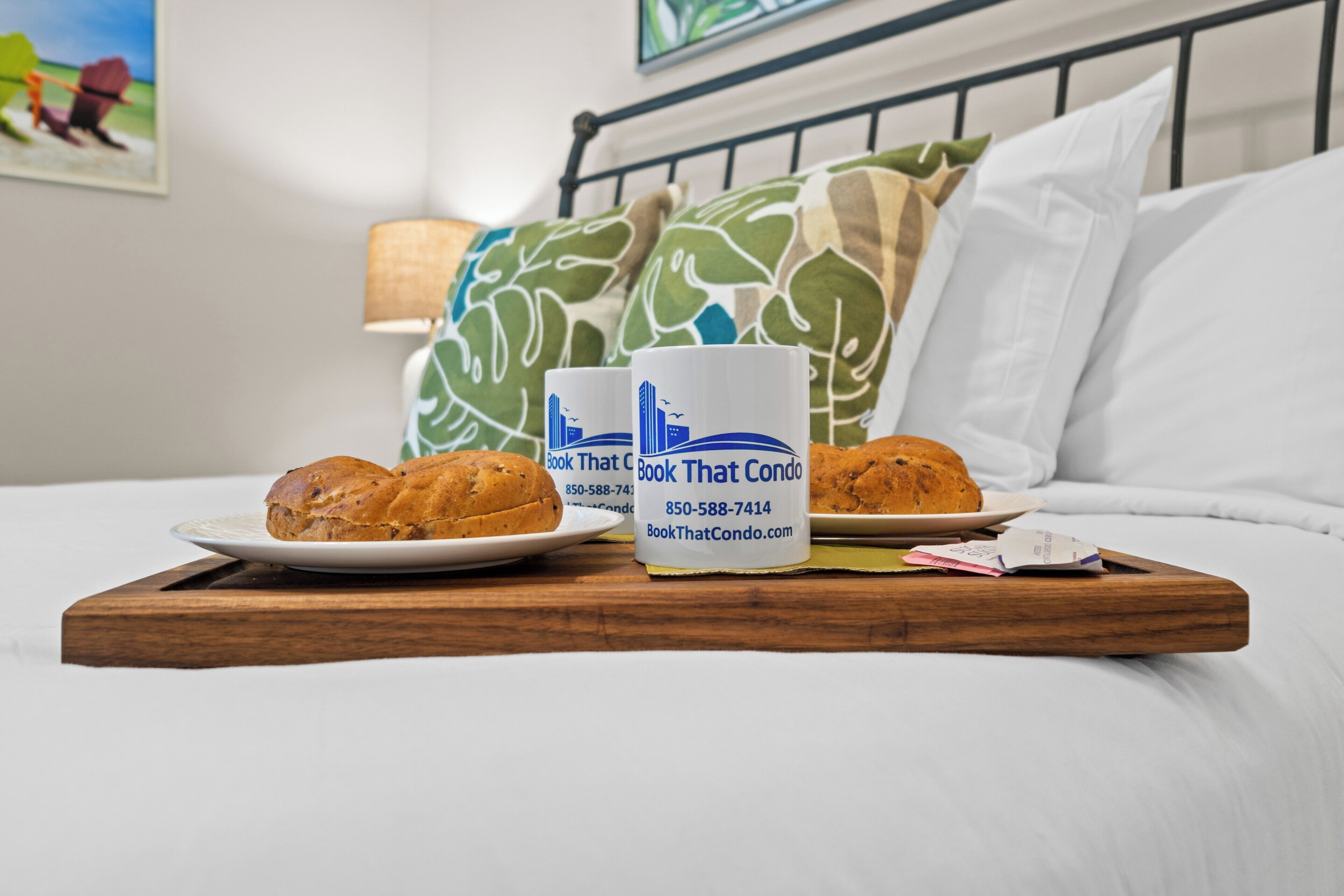 Una cama con dos almohadas, una lámpara de noche y una bandeja con dos muffins y una taza.