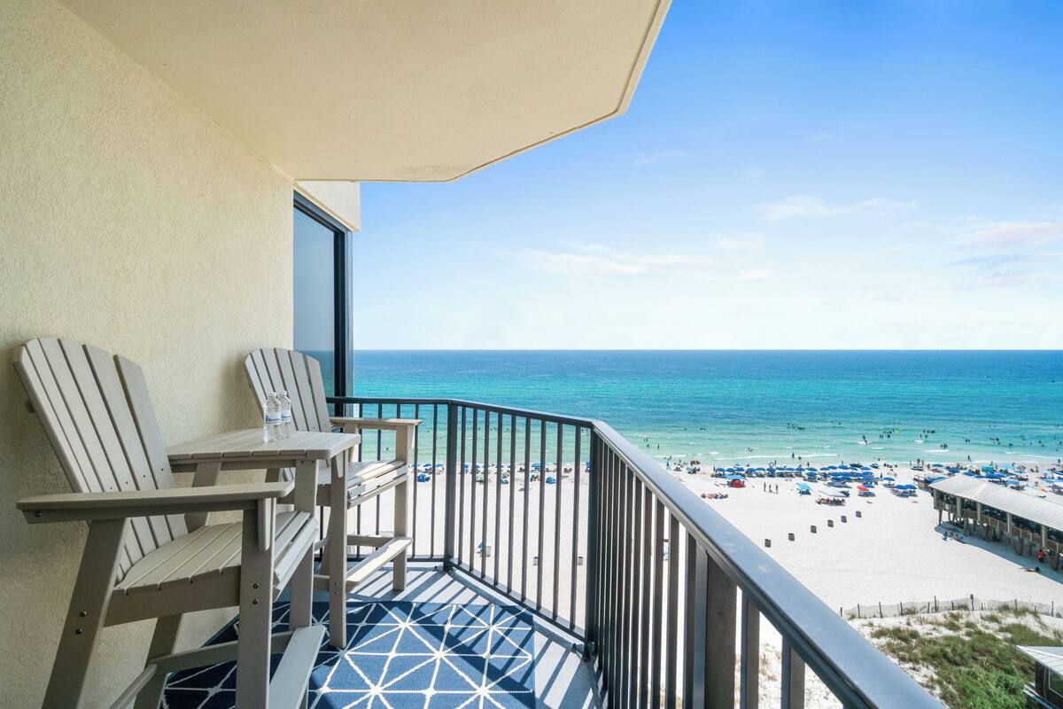 Ein Balkon mit zwei Adirondack-Stühlen, einem Tisch und Blick auf einen Strand mit Sonnenschirmen und Menschen.