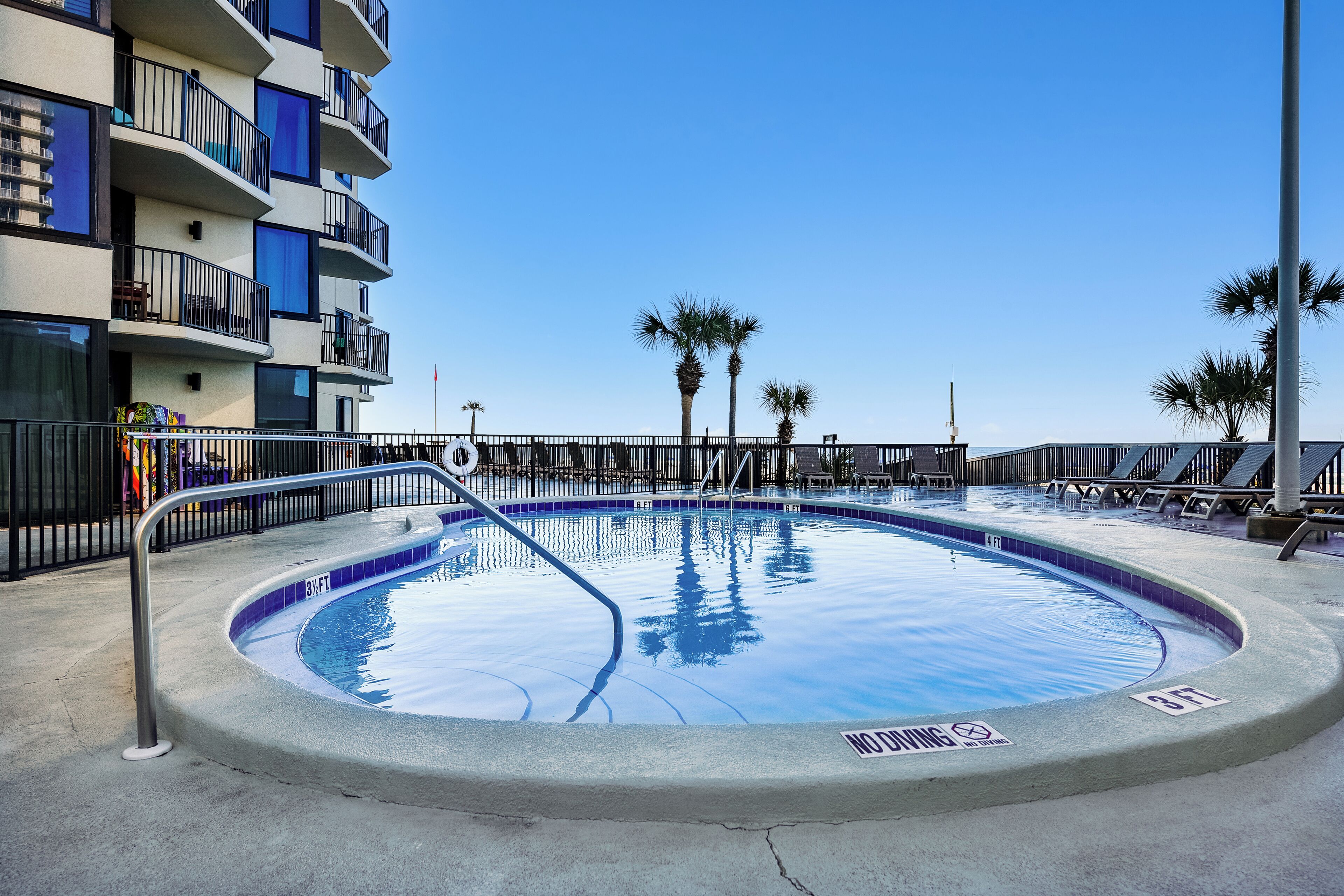 A swimming pool with a no diving sign, surrounded by a fence, with palm trees and a building in the background.
