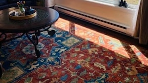 A room with a large window, a dark-colored sofa, a round table with a tray, and a patterned rug.