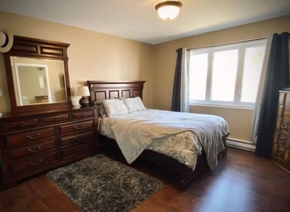 Une chambre à coucher avec une commode en bois, un lit recouvert d’une couvre-lit à motifs, un grand miroir et une fenêtre avec des rideaux.