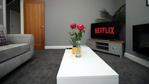 A living room with a grey sofa, a white coffee table, a vase with flowers, a TV with 'NETFLIX' on the screen, and a plant. - Carlton Grove by Tŷ SA (Newport)