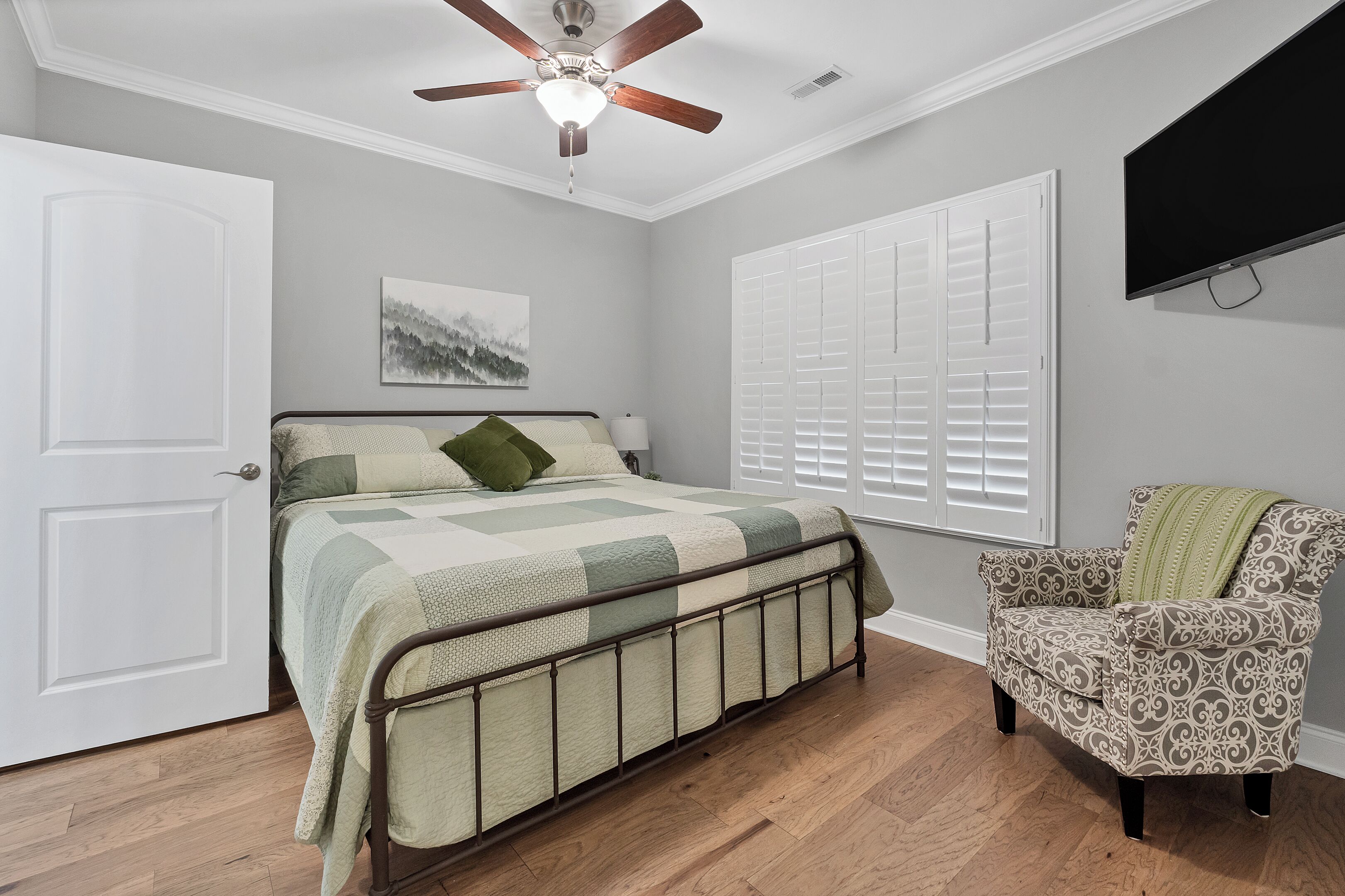 A bedroom with a bed, a chair, a ceiling fan, and a TV.