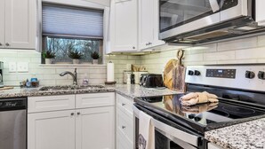 A modern kitchen with white cabinetry, a stainless steel dishwasher, a black stove, and a window with a view of trees.
