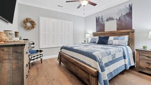 A bedroom with a bed, a dresser, a chair, a ceiling fan, a wall-mounted TV, and a door.