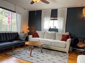 A modern living room with a sofa, a coffee table, and a chair. There is a window with curtains, a ceiling fan, and a floor lamp.