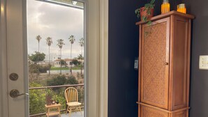 A room with a wooden cabinet against a dark wall, a door leading to a balcony with a chair and a small table, and a view of palm trees and houses.
