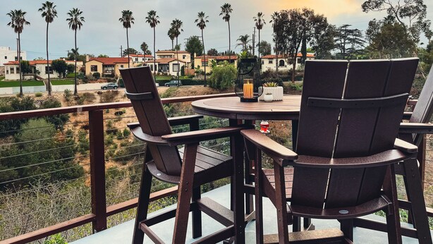 A balcony with a table and chairs, overlooking a residential area with palm trees and houses.