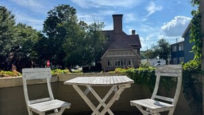 Een terras met twee houten stoelen en een tafel, omgeven door een lage bakstenen muur, met een groot huis en bomen op de achtergrond.