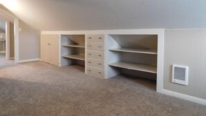 Ein Zimmer mit schrägen Wänden, eingebauten Stauraum mit mehreren Schubladen und Regalen, einem Teppichboden und einem Lüftungsgitter an der Wand.