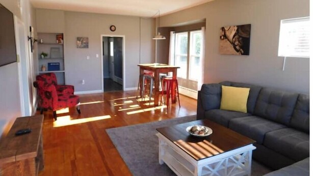 Ein geräumiges Wohnzimmer mit grauem Sofa, einem Couchtisch und einem roten Sessel. Es gibt eine Essecke mit Tisch und Stühlen.