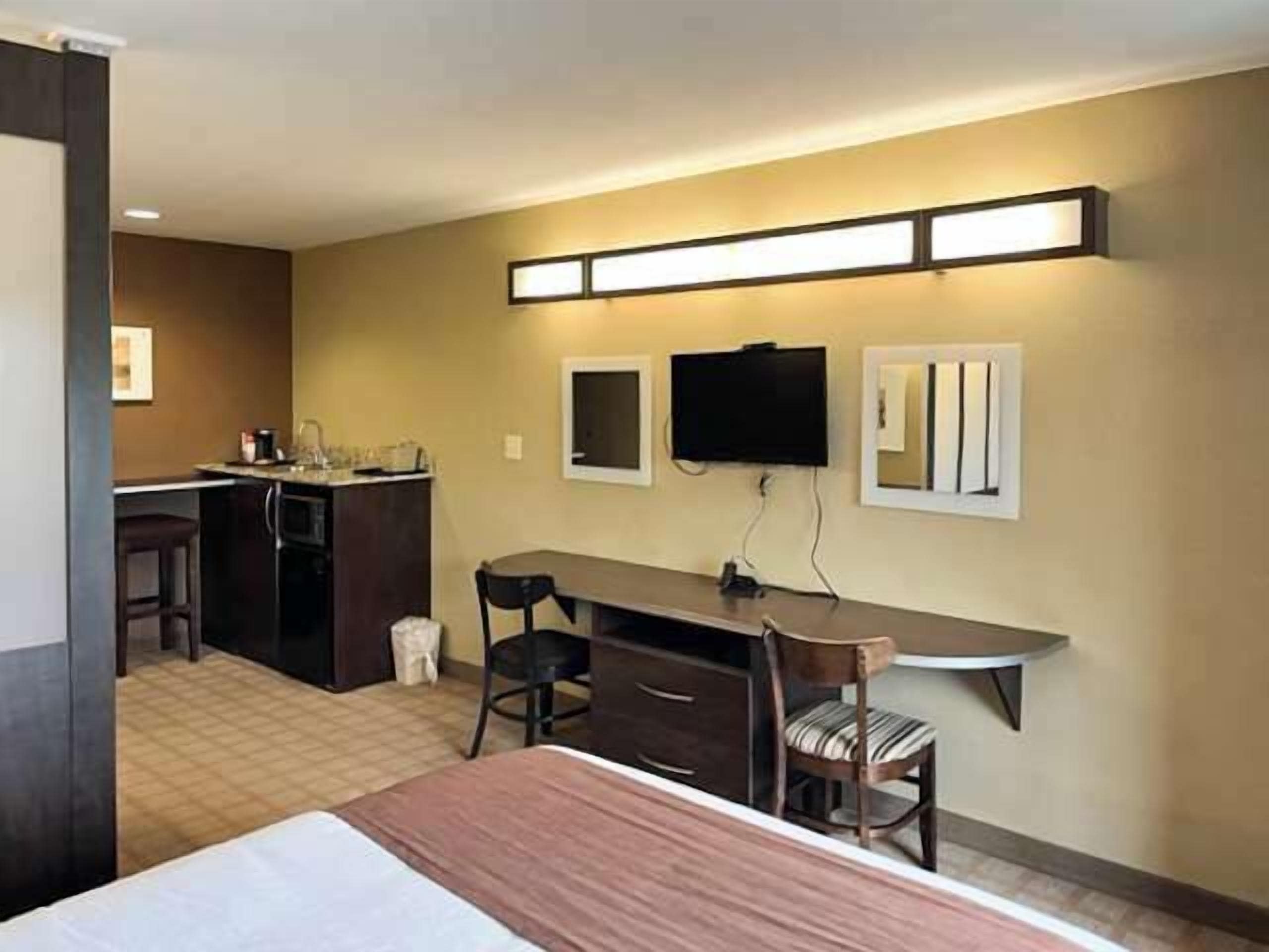 A hotel room with a bed, a desk, a chair, a TV, and a kitchenette.