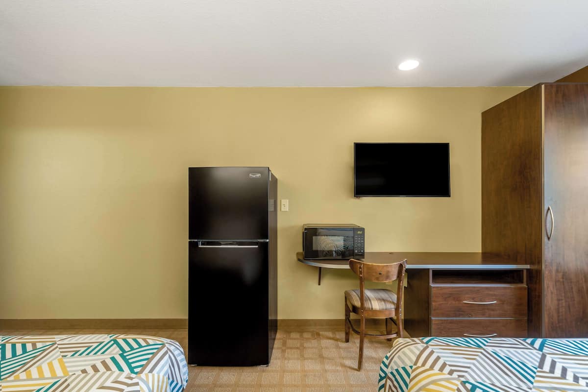 A compact hotel room with a bed, a refrigerator, a microwave, a small table, and a TV mounted on the wall.