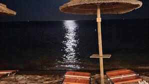Una spiaggia con ombrelloni di paglia e lettini, un palo di legno e un mare calmo sotto un cielo notturno con una luna visibile.