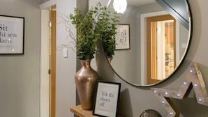 A hallway with a round mirror, a framed sign saying "Tek thi shoes off", and a potted plant on a shelf. - Remarkable 4-bed House Near Leeds Airport (Leeds)