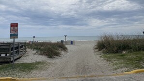Uma trilha de areia que leva ao oceano, com uma placa de 'Não Estacionar' e uma torre de salva-vidas ao longe.