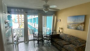 Sala de estar com mesa de vidro, duas cadeiras, um sofá, ventilador de teto e vista para um prédio azul lá fora.