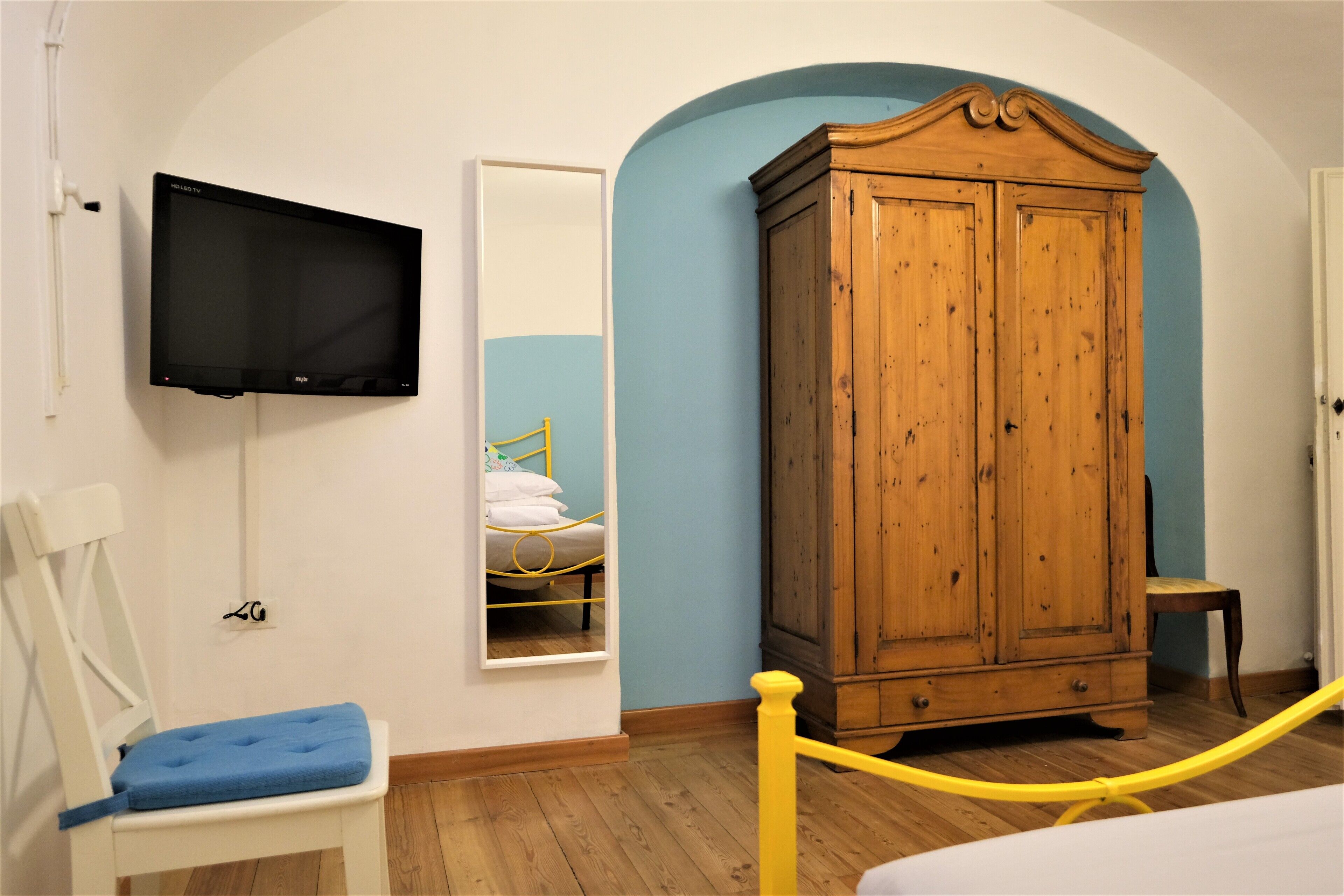 A room with a wooden wardrobe, a television mounted on the wall, a chair with a blue cushion, and a yellow rope barrier.