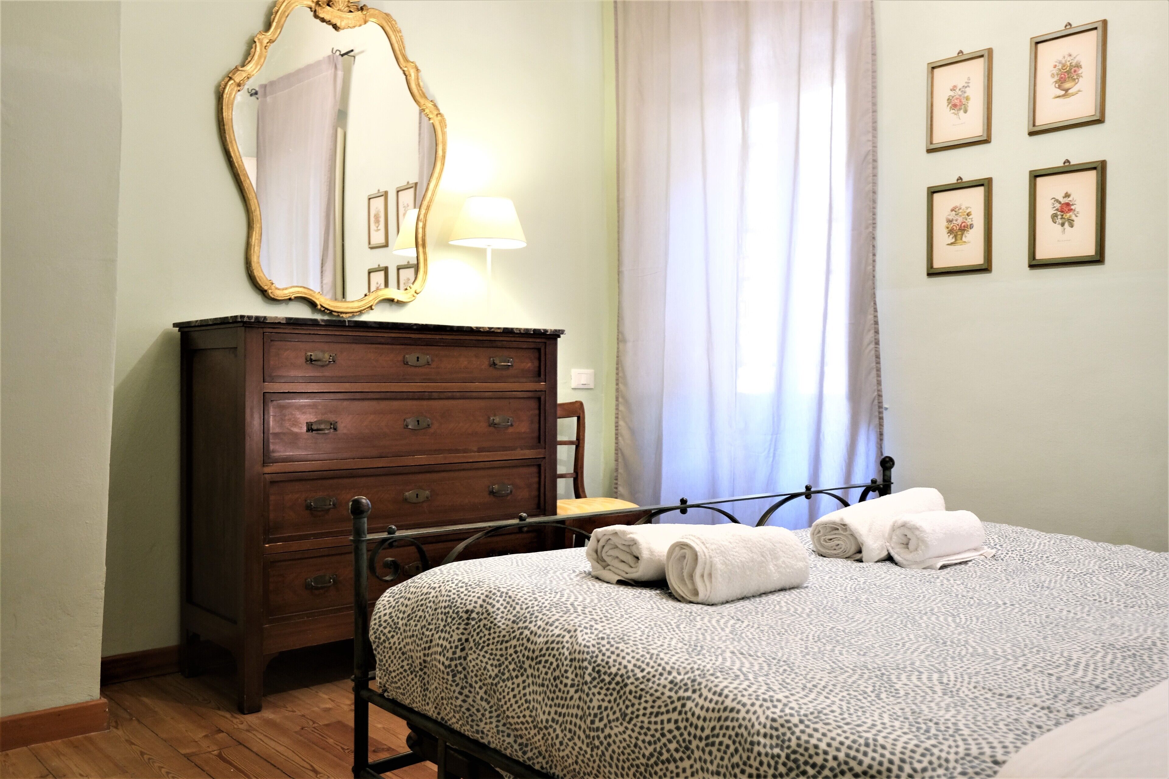 A bedroom with a wooden dresser, a bed with a patterned bedspread, a mirror with a gold frame, a lamp, and four framed floral pictures on the wall.