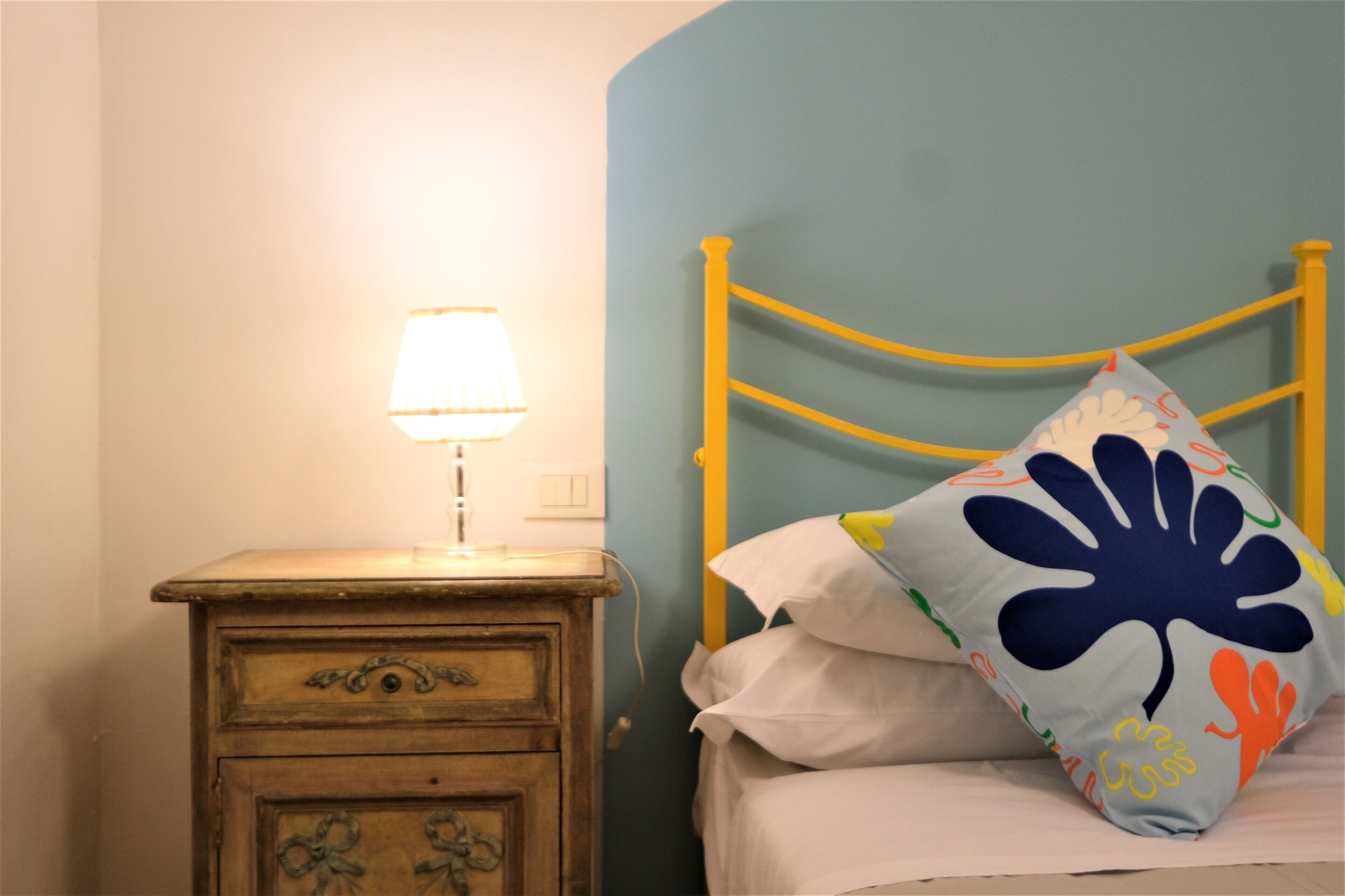 Un dormitorio con una mesita de noche de madera, una lámpara, una cama con un cojín colorido y una pared azul.