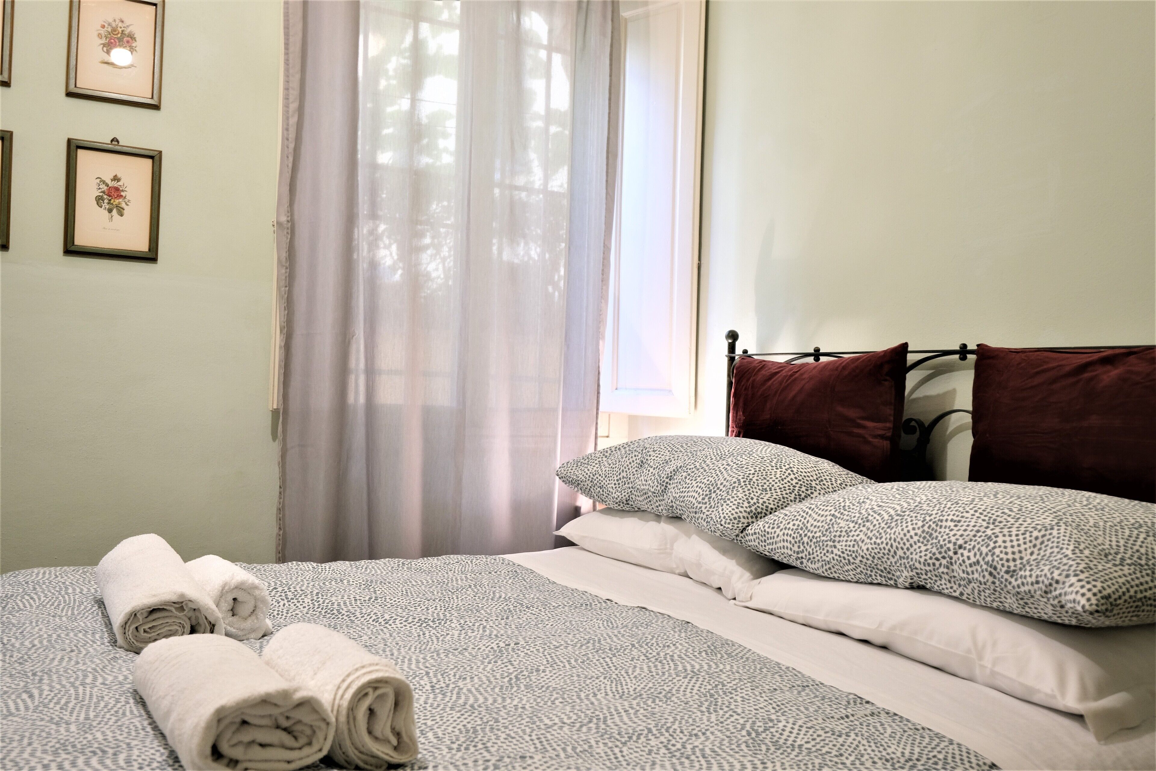 A neatly made bed with rolled towels, a window with sheer curtains, and framed floral artwork on the wall.