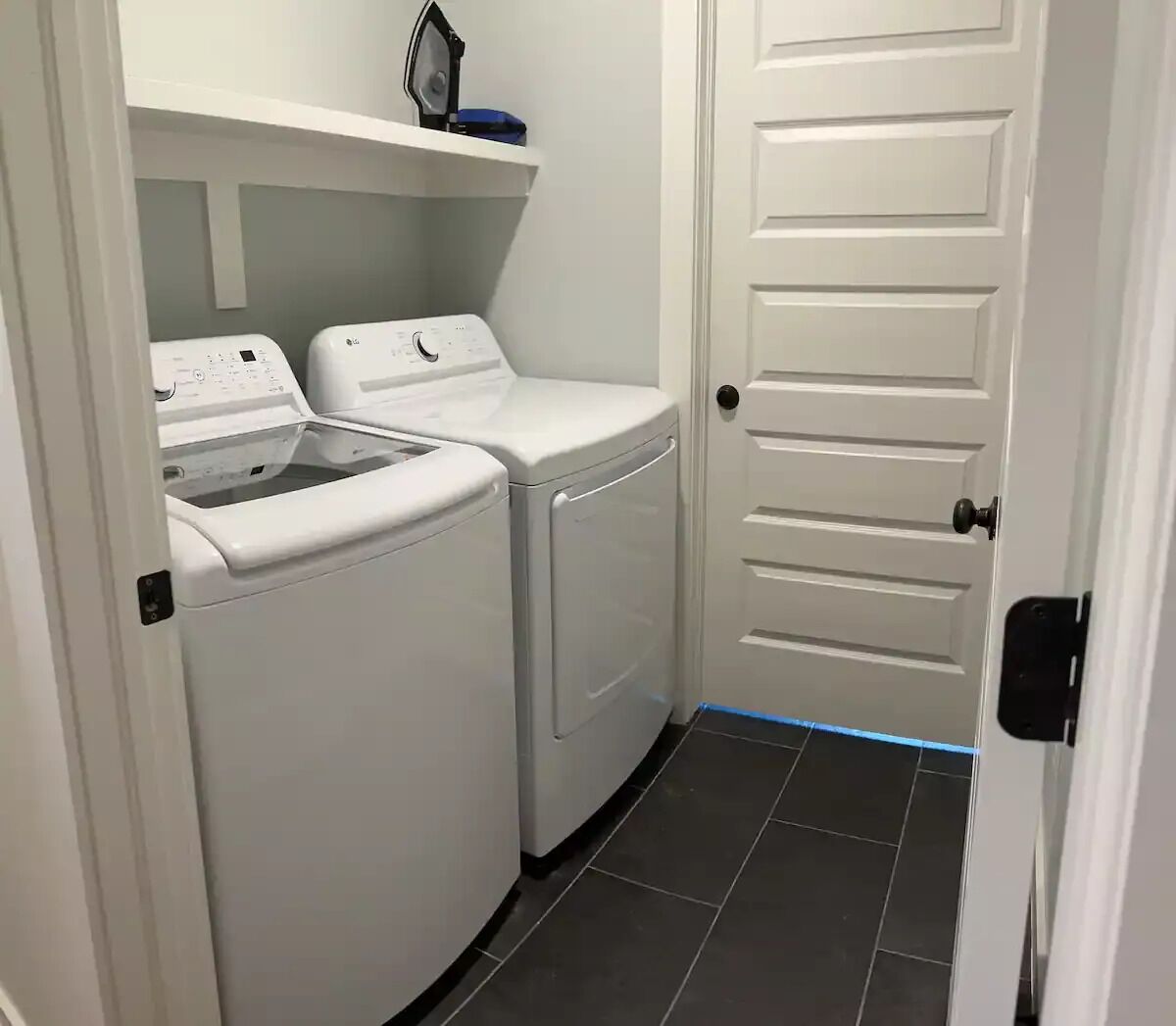 A laundry room with two white washing machines and a dryer, a shelf with an iron, and a white door.