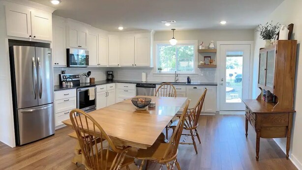 A modern kitchen with white cabinets, stainless steel appliances, a wooden dining table, and a large window. - The Red Gate - Branson & Eureka!
 (Green Forest)