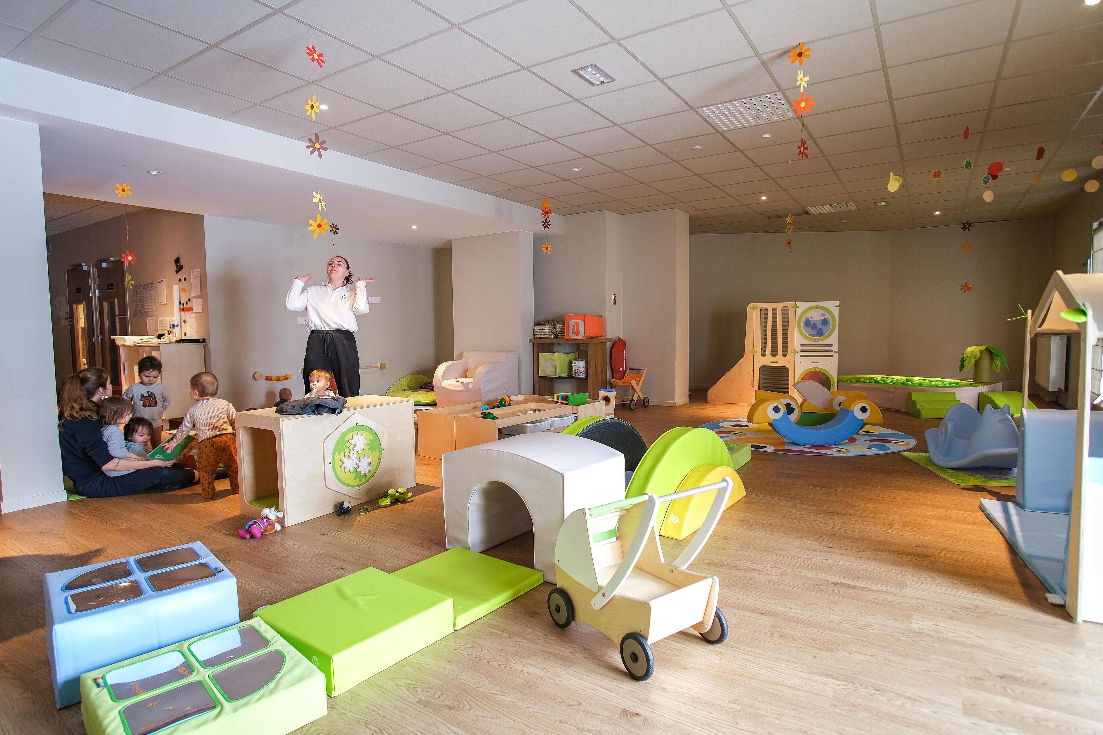 Uma sala de jogos infantil com diversas estruturas de recreação, um espaço de cozinha e uma área de estar com adultos supervisionando as crianças.
