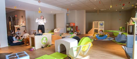 A children's playroom with various play structures, a kitchen area, and a seating area with adults supervising children.