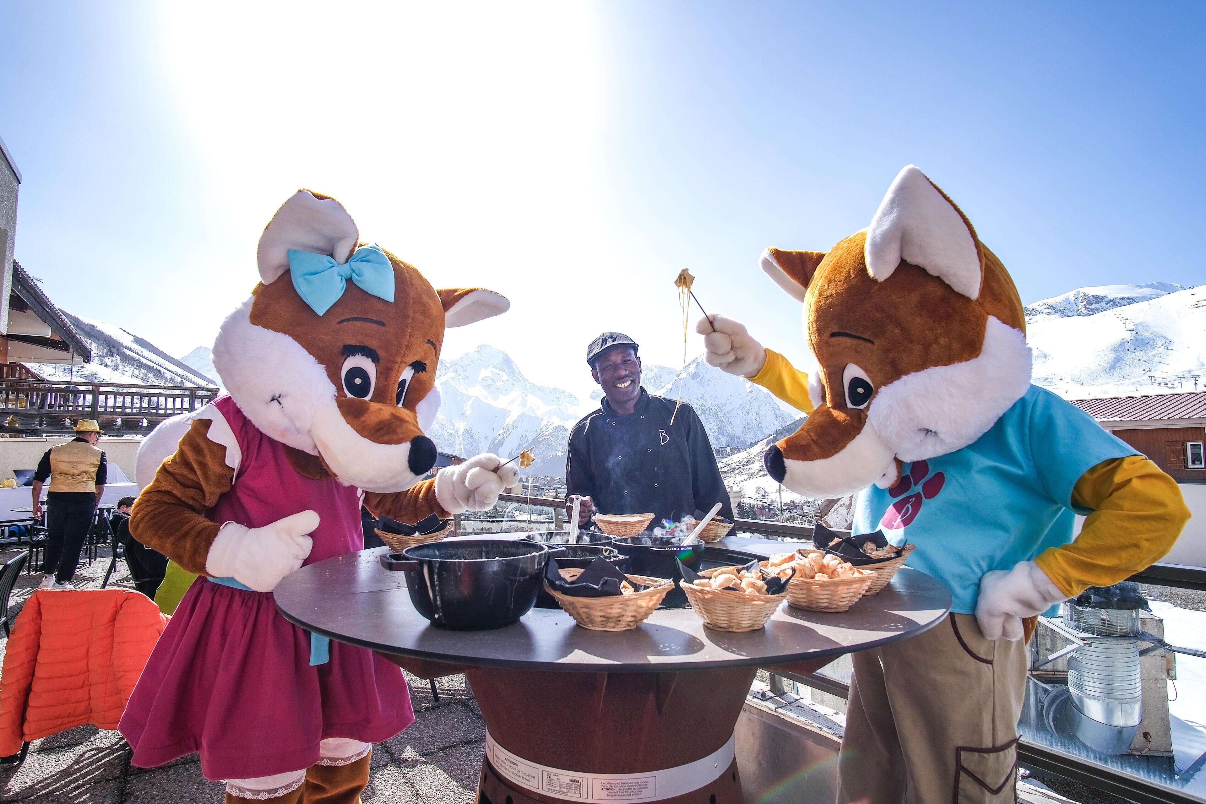 Due personaggi pubblicitari servono del cibo a un tavolo con montagne sullo sfondo.