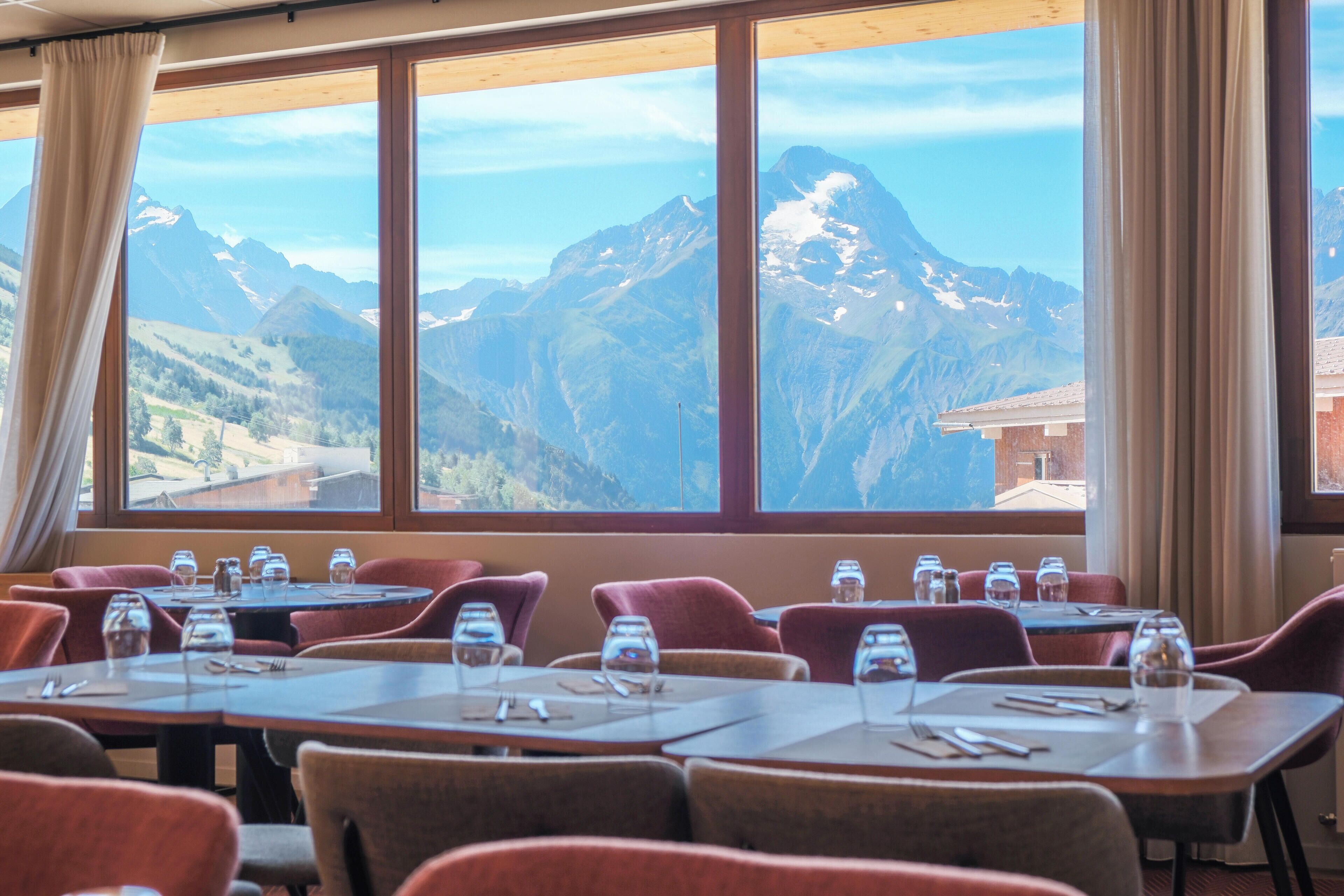 Un restaurante con mesas dispuestas para comer, amplias ventanas con vistas a las montañas y sillas tapizadas en rojo.