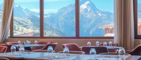 Un restaurante con mesas dispuestas para comer, amplias ventanas con vistas a las montañas y sillas tapizadas en rojo.