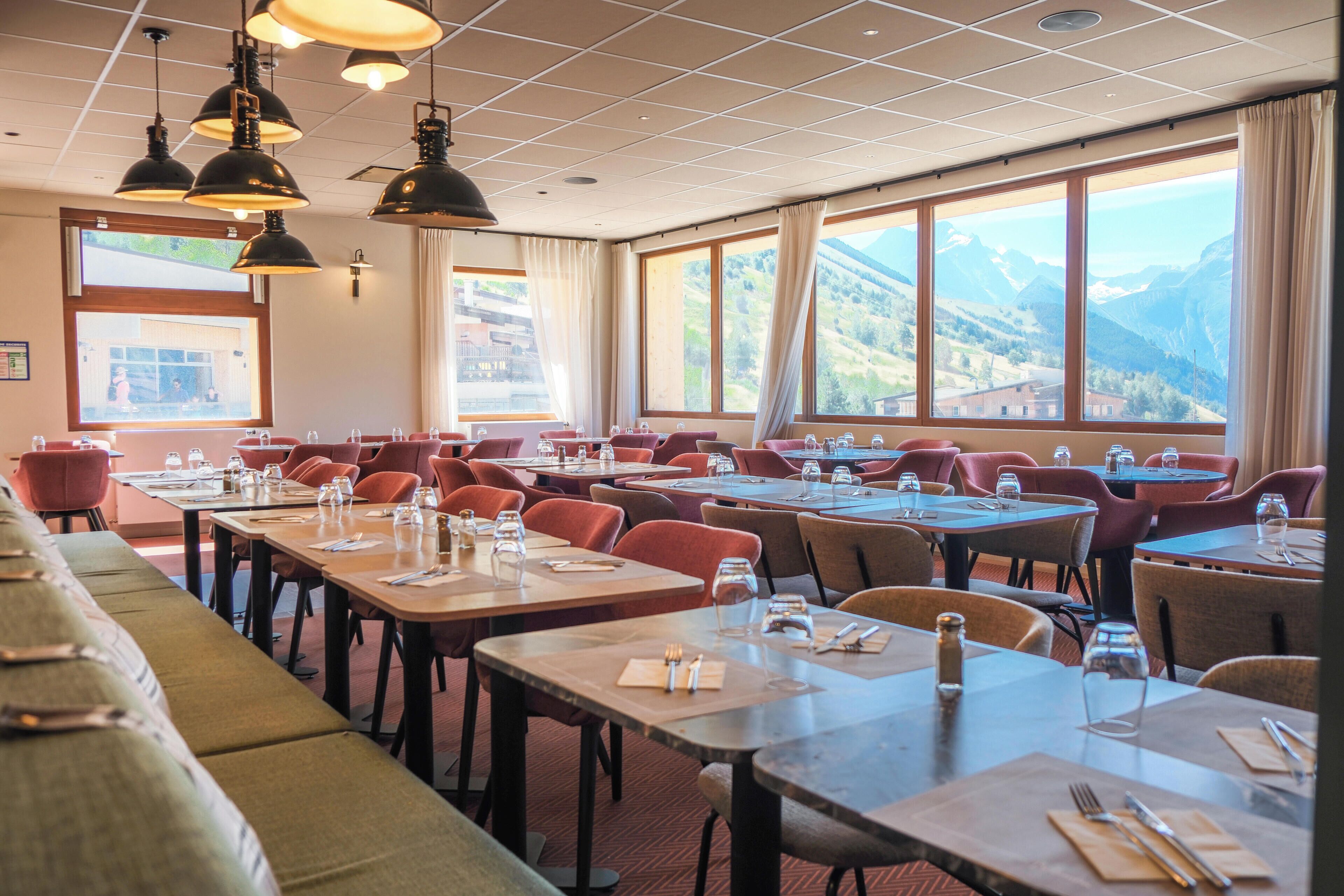 Un restaurante con varias mesas y sillas, amplias ventanas con vistas a la montaña y lámparas colgantes.
