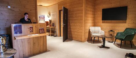 A wooden interior with a reception desk, a seating area with a chair and a table, and a mounted screen on the wall.